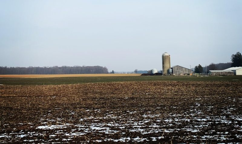 Snow and soil in the foreground of a picture which leads though a farm field, past a silo and barn, and into the blue sky.