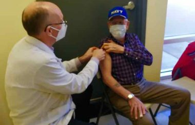 A male pharmacist wearing a white jacket and white mask gives a man a COVID-19 vaccine at a clinic. The man receiving the vaccine has his plaid shirt sleeve rolled up and he's wearing a blue hat and face mask.