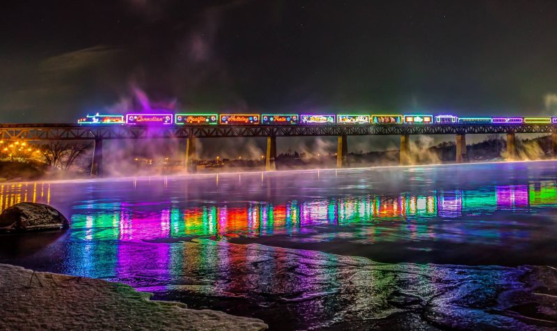The CP Holiday Train lit up in festive colours passing over a lake.