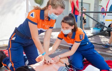 Two women in orange and blue uniform are practicing CPR on a mannequin.