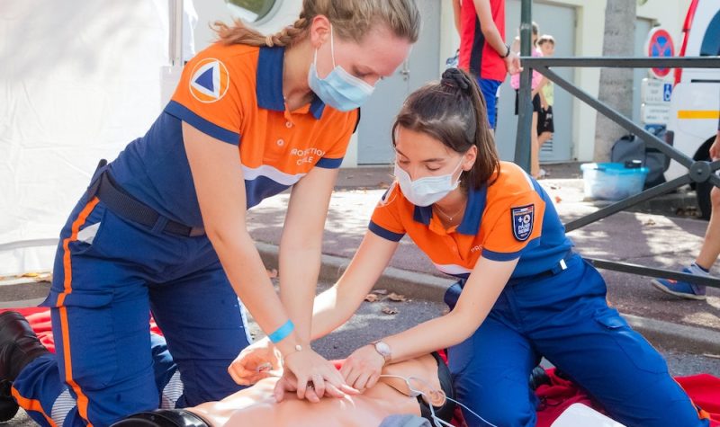 Two women in orange and blue uniform are practicing CPR on a mannequin.