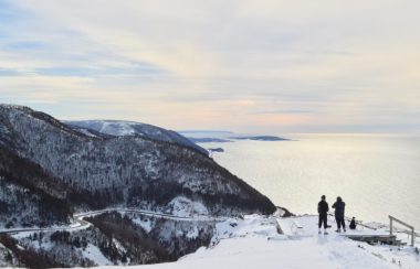 Deux personnes au bord d'une falaise regardant la route et les montagnes enneigées.