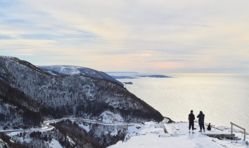 Deux personnes au bord d'une falaise regardant la route et les montagnes enneigées.