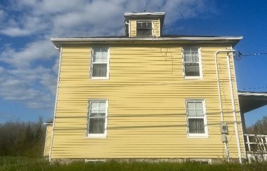 A yellow two-storey house sits on a grassy lot with trees in the background.