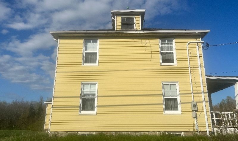 A yellow two-storey house sits on a grassy lot with trees in the background.