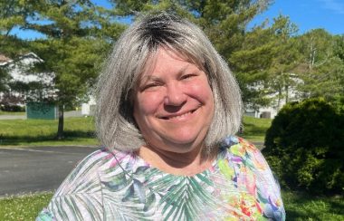 A woman with grey hair wearing a colourful printed shirt smiles against a background of a concrete road, trees and a blue sky.