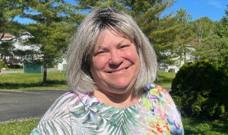 A woman with grey hair wearing a colourful printed shirt smiles against a background of a concrete road, trees and a blue sky.