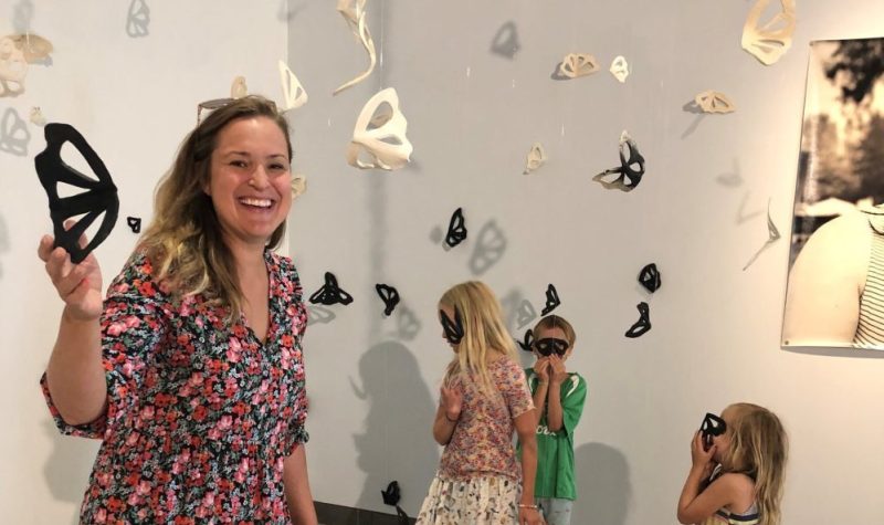 On peut voit l'artiste Camille Myles et ses trois enfants qui sont photographiés en plein coeur de l'exposition « Émergence » réalisée par Myles. On aperçoit des papillons noirs et blancs fait en céramique, ainsi qu'une partie de portrait faisant partie de l'exposition. La pièce où l'exposition à lieu a des murs blancs.