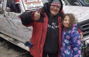 A middle-aged woman wearing a red jacket over a black hoodie with a grey toque stands with her arm around a young girl wearing a purple jacket, smiling at the camera and giving a thumbs up. A white vehicle is seen behind them.