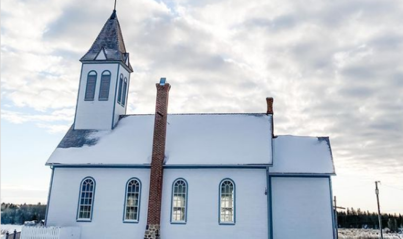 Une petite église blanche est recouverte de neige. Au centre du bâtiment se trouve une cheminée en brique rouge. Deux fenêtres longues sont de chaque côté de la cheminée. Le sol est couvert de neige. Le ciel est nuageux et blanc.