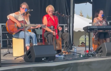 Trois femmes sont assises sur une scène. L'une est à la guitare, l'autre au clavier et la troisième est assise sur une caisse de percussion. Les trois femmes ont un micro devant elles.