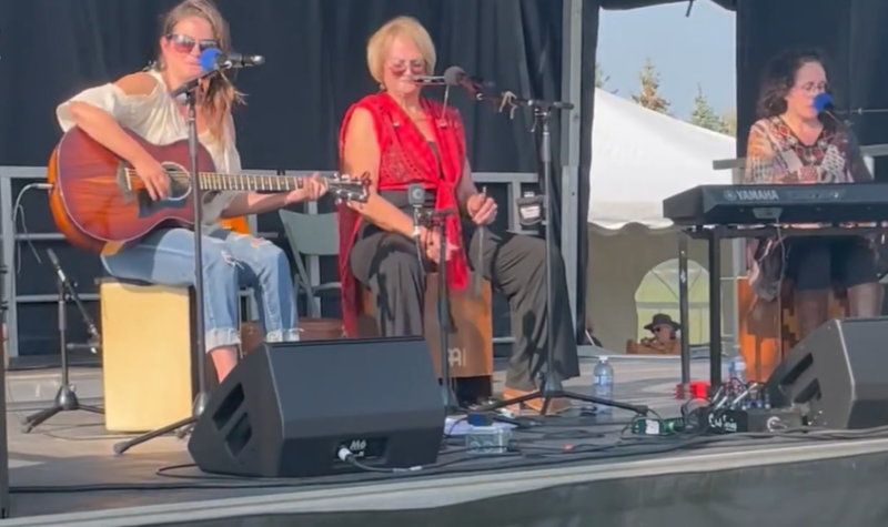 Trois femmes sont assises sur une scène. L'une est à la guitare, l'autre au clavier et la troisième est assise sur une caisse de percussion. Les trois femmes ont un micro devant elles.