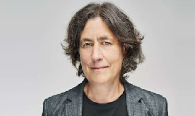 Une femme habillée d'un veston gris souris à la caméra en plan rapproché.