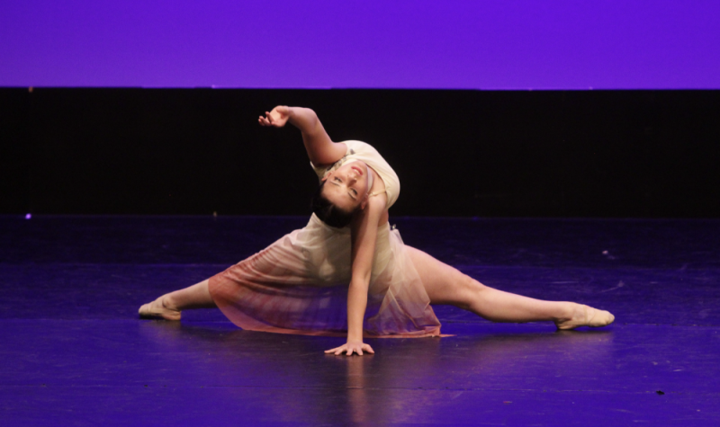 Une jeune fille fait le grand écart. Elle a la tête renversée, une main la supportant au sol derrière elle et une main dans les airs. Elle est sur une scène bleue. Elle porte une robe de ballerine.