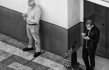 En noir et blanc, deux personnes, un homme grisonnant et un d'une trentaine d'années portent un masque et se tiennent à distance. Les deux consultent leurs téléphones.
