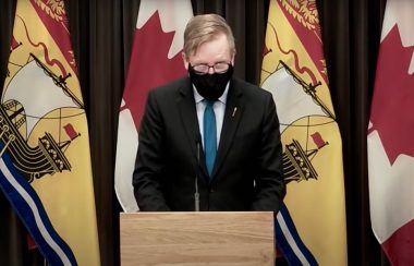 New Brunswick Education Minister Dominic Cardy speaks at a press conference on Dec. 31, 2021. He is standing at a podium, wearing a black face mask and a suit, in front of several flags.