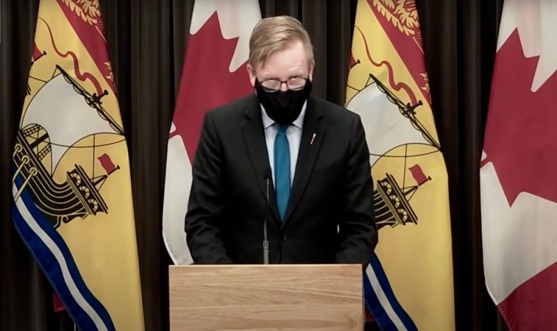 New Brunswick Education Minister Dominic Cardy speaks at a press conference on Dec. 31, 2021. He is standing at a podium, wearing a black face mask and a suit, in front of several flags.