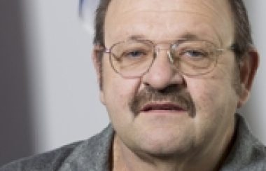 A professional headshot of Carl Mayer, wearing glasses and a grey golf shirt in front of a Quebec flag.