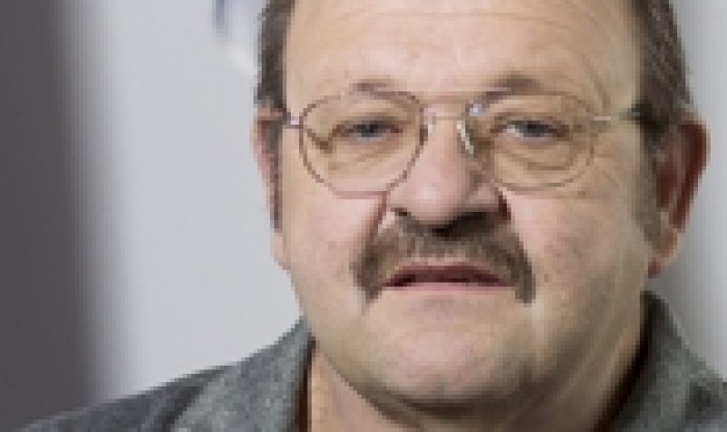 A professional headshot of Carl Mayer, wearing glasses and a grey golf shirt in front of a Quebec flag.