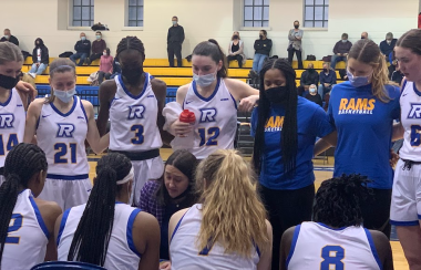 Basketball players in white jerseys stand in a circle around a person kneeing in the middle.