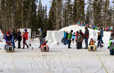 Des enfants font une course de tracteurs en bois. entourés de leurs parents. On peut voir dehors des sculptures de neige.