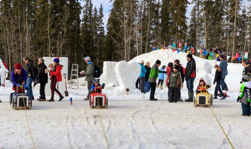 Des enfants font une course de tracteurs en bois. entourés de leurs parents. On peut voir dehors des sculptures de neige.