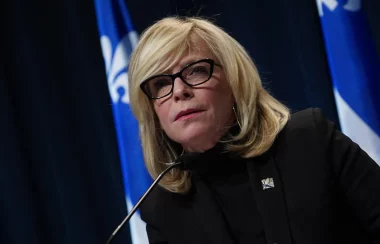 La ministre Caroline Proulx vêtue d'un veston noir, penchée vers l'avant. Sur la toile de fond, deux drapeaux du Québec.