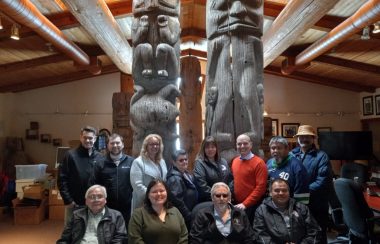 The Gitanyow Band Chiefs and Executive Director Joel Starlund along with Stikine MLA Nathan Cullen pose for a photo in front of two looming totem poles on Gitanyow Territory.