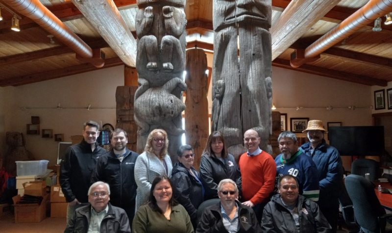 The Gitanyow Band Chiefs and Executive Director Joel Starlund along with Stikine MLA Nathan Cullen pose for a photo in front of two looming totem poles on Gitanyow Territory.