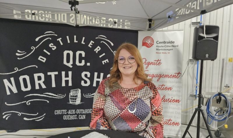 Une jeune femme avec les cheveux roux et lunettes debout devant des affiches représentant une distillerie