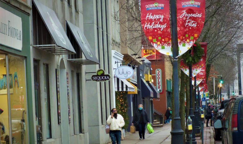 Un lampadaire avec des affiches souhaitant un joyeux Noël en français et en anglais; une femme parlant au téléphone marche sur le trottoir; plusieurs commerces sont à sa droite