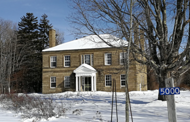 A two story stone house, with snow covered landscape in foreground and address marker reading 5000.