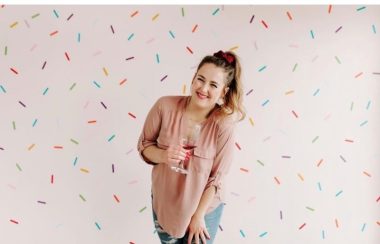 A photo of Bon Apatreat bakery owner Chantelle Villeneuve. Sh is centered in the photo, wearing a pinkish tshirt, blue jeans, a red bow in her high pony tail which matches her lipstick and the wine in the glass she is holding. The background is all confetti.