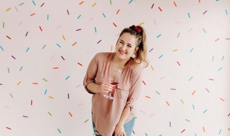 A photo of Bon Apatreat bakery owner Chantelle Villeneuve. Sh is centered in the photo, wearing a pinkish tshirt, blue jeans, a red bow in her high pony tail which matches her lipstick and the wine in the glass she is holding. The background is all confetti.