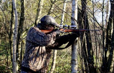 Parmi des arbres dans une forêt, un homme de côté vise à l'extérieur de la photo une cible avec une carabine.