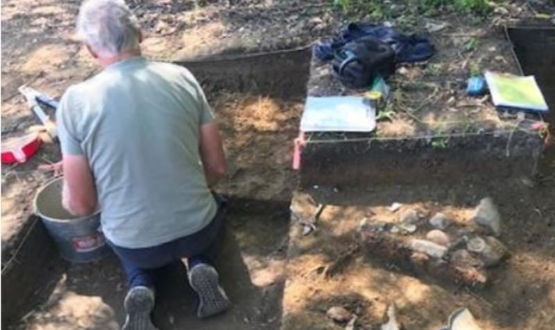 A man kneels with his back to the camera in a rectangular hole in the dirt at an archaeological dig site. There is a bucket in front of him and digging tools next to him on the ground to the right of the hole.