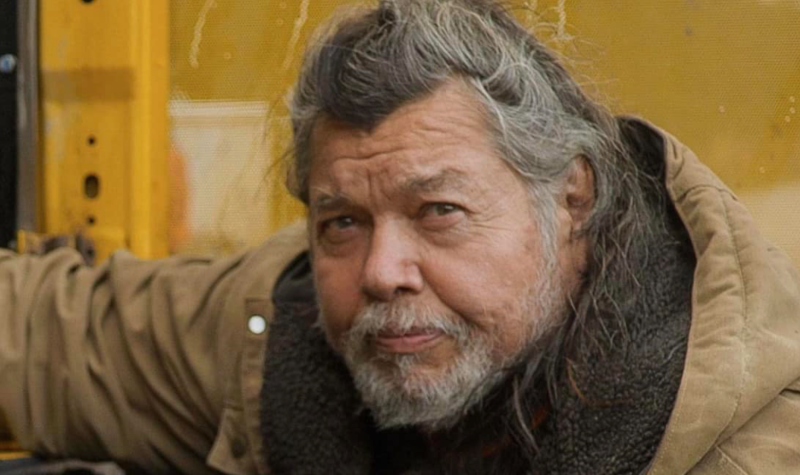 A bearded man with greying hair sits in front of a yellow coloured wall. He is wearing a light brown winter coat.