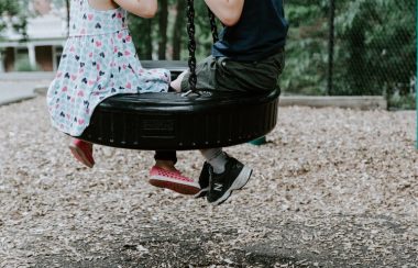 Children's legs dangle off of a tire swing as they sit on it, with a schoolyard in the background.