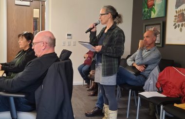 A woman wearing a plaid jacket and white boots stands at a council meeting with a mic in her hand. There are four people sitting beside and in front of her.
