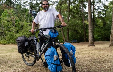 A man wearing sunglasses and a white shirt poses with a mountain bike with several carrier bags in Bowen Park in Nanaimo.