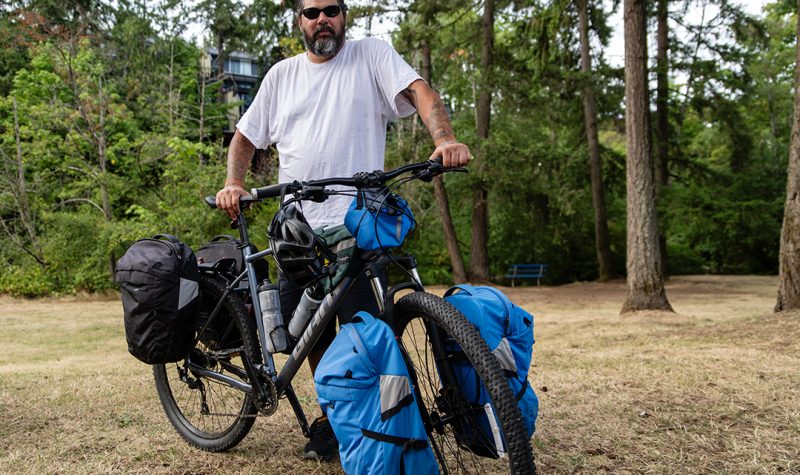 A man wearing sunglasses and a white shirt poses with a mountain bike with several carrier bags in Bowen Park in Nanaimo.