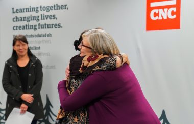Two people embrace each other while another looks on in front of a white, red and grey background with the lettering 'CNC' and 'Learning together, changing lives, creating futures' on the banner.