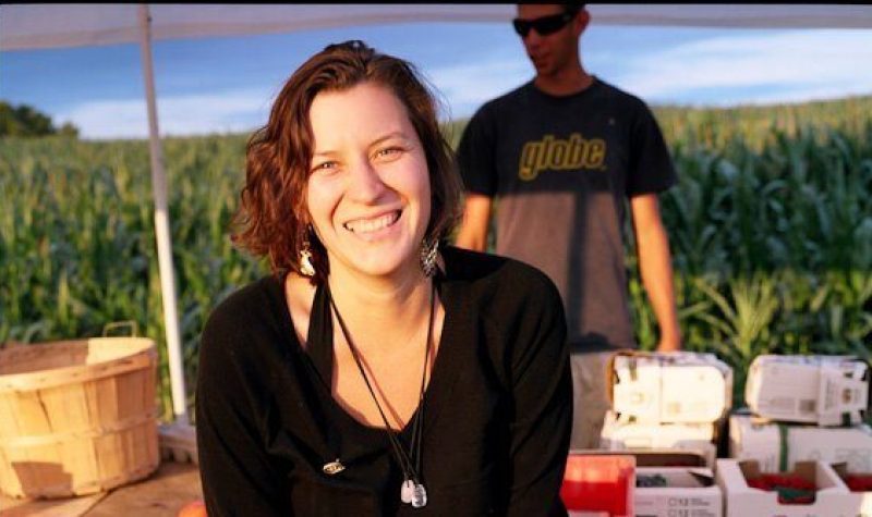 A woman with brunette hair dressed in black and smiling is at an outdoor event with a blue sky and clouds on the horizon.