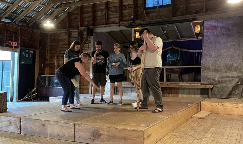 Seven actors stand on a wooden stage under a gabled roof. There is a small window behind the stage and open door beside it.