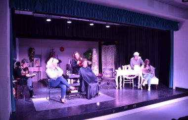 A group of 6 women on stage performing the play Steel Magnolias, with their scene set in a hairdresser's shop.