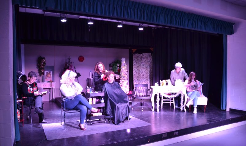 A group of 6 women on stage performing the play Steel Magnolias, with their scene set in a hairdresser's shop.