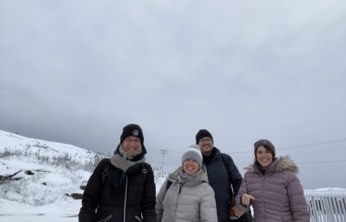 Quatre personnes en manteaux d'hiver sourient joyeusement à la caméra devant un paysage brut et enneigé.