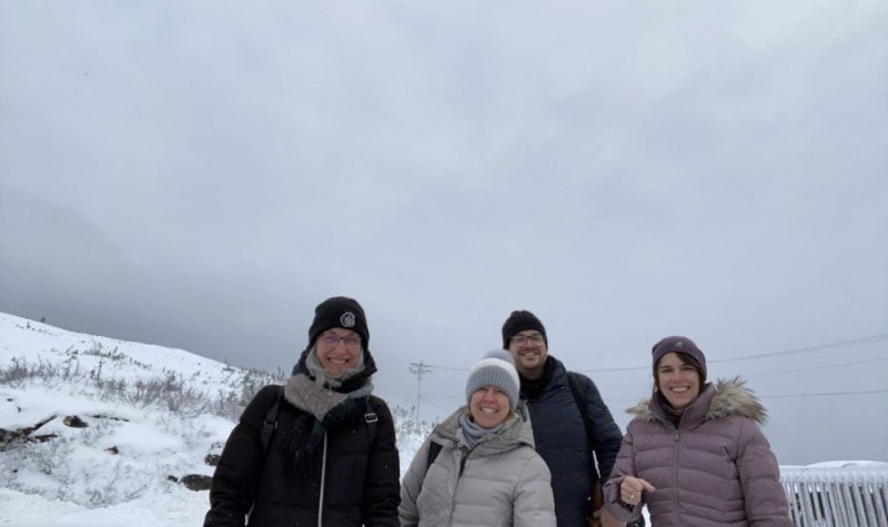 Quatre personnes en manteaux d'hiver sourient joyeusement à la caméra devant un paysage brut et enneigé.