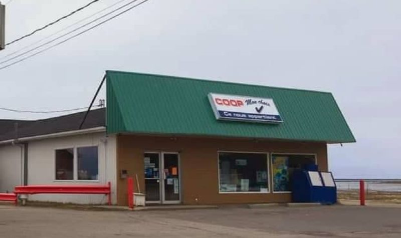 Magasin alimentaire avec devant vert et beige, le côté de la bâtisse est blanc et il y a un stationnement en asphalte devant et sur le côté. On voit un ciel dégagé et la mer derrière.