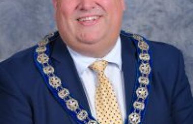 Corner Brook Mayor Jim Parsons is smiling and wearing a blue jacket and yellow tie. He is also wearing the chain of office around his neck.